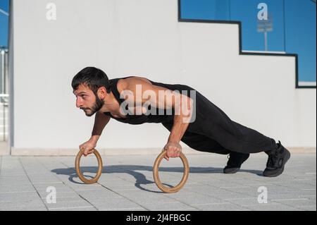 A man in black sportswear doing ring push-ups outdoors. Stock Photo