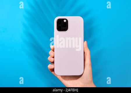 Female hand holding white smartphone in soft silicone cover back view . Phone case mock up isolated on blue background shadow from a plant Stock Photo