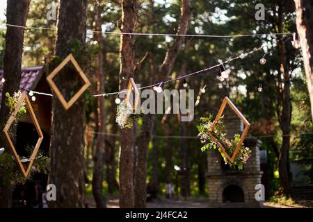 Wedding party banquet outdoors in pine forest. Dining tables, benches decorated in boho style with candles, flowers, spring fairy lights, plates Stock Photo