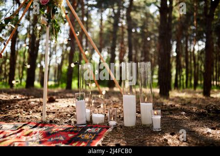 Candles burning in glass vases, flasks on carpet near bohemian tipi arch decorated with roses and flowers wrapped in fairy lights on outdoor wedding Stock Photo