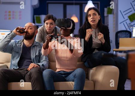 African american woman using vr glasses and playing video games at office party with drinks after work hours. Colleagues having fun with virtual reality headset at celebration. Stock Photo