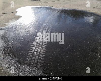 Track from a studded tire on the ice of a frozen puddle on cracked asphalt in the bright sunshine on a winter day. Stock Photo