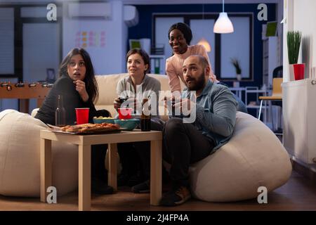 Smiling people playing video games online on tv console, using controller to win at office part after work. Colleagues having fun with game, eating pizza and drinking alcoholic beer. Stock Photo