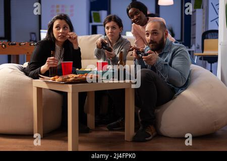 Diverse group of people playing video games on console, using controller on tv at office celebration with drinks and food. Colleagues enjoying after work party with alcoholic beverage and game. Stock Photo