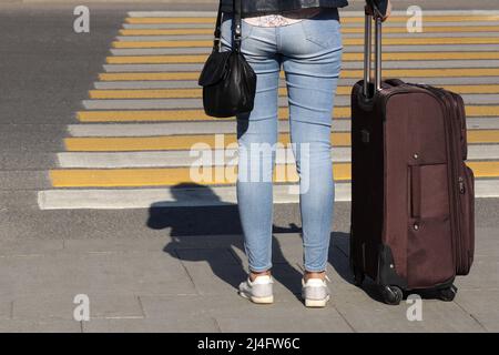 Woman in jeans stands with suitcase on wheels on a pedestrian crossing, rear view. Female legs in front of crosswalk, concept of travel, waiting taxi Stock Photo