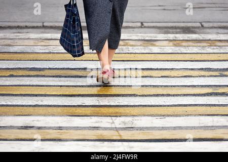 Old woman in long skirt with checkered bag walks on pedestrian crossing. Road zebra marking, street safety concept Stock Photo