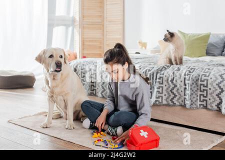 preteen girl playing doctor with toy medical instruments near labrador and cat in bedroom Stock Photo