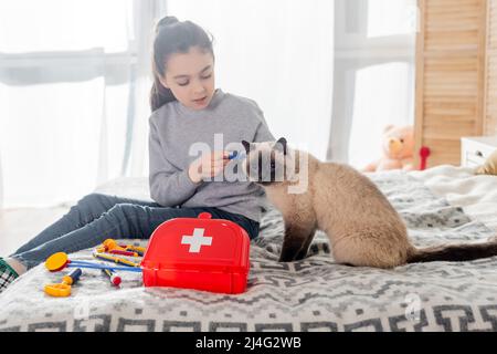 girl making injection to cat with toy syringe near first aid kit on bed Stock Photo