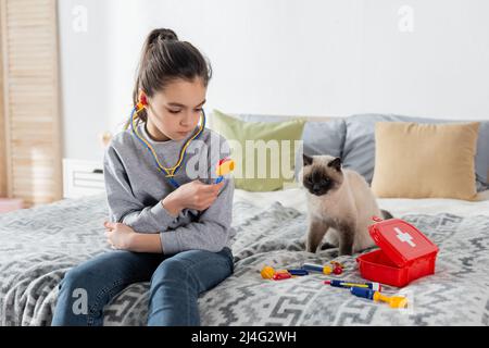 girl holding toy stethoscope while playing on bed near cat Stock Photo