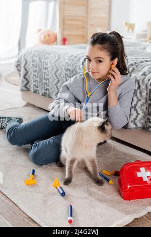 happy girl smiling at camera while examining cat with toy stethoscope on floor Stock Photo