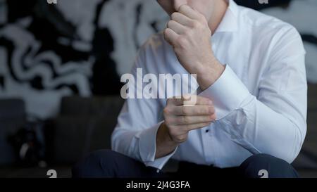 The man gets dressed and does his buttons on the sleeves of his white shirt. Action. Close up of male arms, man sitting and trying to button up. Stock Photo