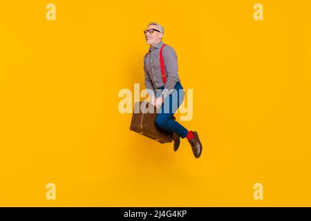 Full body photo of funny grey haired pensioner flying around world on old retro bag isolated on yellow color background Stock Photo