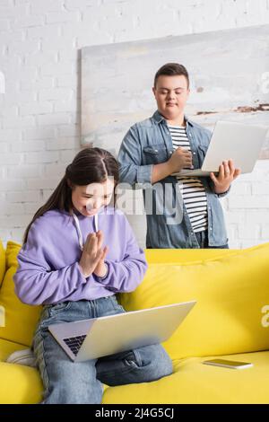 Cheerful teenager with down syndrome using laptop near smartphone and friend at home Stock Photo