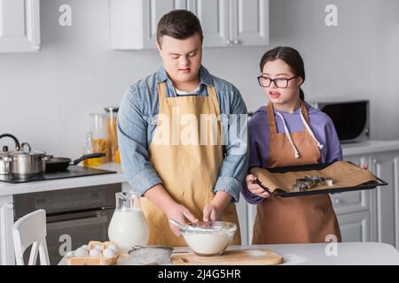 Teen couple with down syndrome in aprons cooking in kitchen Stock Photo