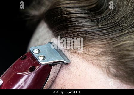 Young bearded man with a mohawk getting haircut by hairdresser while sitting in chair at barbershop and smoking a cigarette Stock Photo