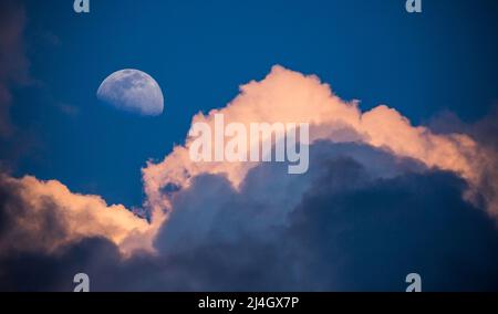 A waxing gibbous moon rises in a deep blue sky above gathering storm clouds lit by the setting sun. Stock Photo