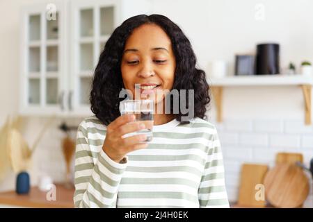 Smiling African-American female enjoying glass of clear water while standing in kitchen Stock Photo