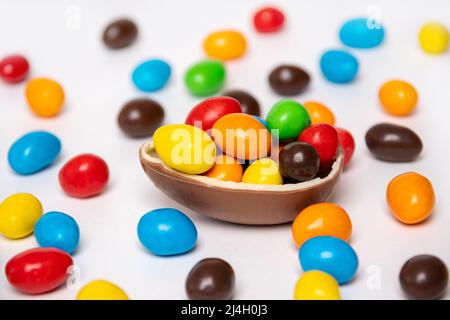 Half of a cracked chocolate egg filled with multi-colored candies on a white background with multi-colored candies. Treats for children. Sweet dessert Stock Photo