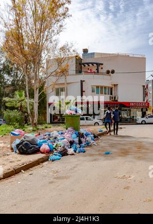 Full dumpster with scaterred garbage bin on the sidewalk. People walking near dustbin bags, boxes and trash can near the Le Carre patisserie. Stock Photo