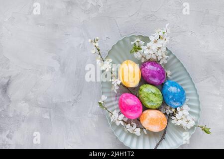 Colorful Easter eggs on plate and spring white cherry blossom flowers on gray background. Easter celebration concept. Top view, flat lay, copy space Stock Photo