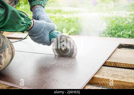 hands of a locksmith in special clothes works in the production of home repairs. Processing of ceramics with an angle grinder. Cuts ceramic tiles with Stock Photo