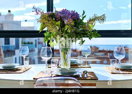 Beautiful table place arrangement with bouquet of flowers in vase, served places with dishware, sea and houses rooftops view through window Stock Photo