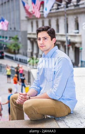 Clamping his hands, a young handsome guy is sitting outside on a stage into deeply thinking. There are American flags hanging on buildings in the back Stock Photo