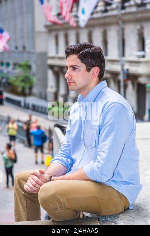 Clamping his hands, a young handsome guy is sitting outside on a stage into deeply thinking. There are American flags hanging on buildings in the back Stock Photo