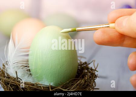 Close-up female hand, a human painting Easter eggs with green paint. Easter egg, white feather in bird's nest. Spring season holiday decoration. Drawi Stock Photo