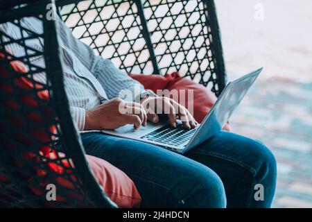 A man is sitting on a hanging rope swing and working on a laptop. Male hands on the keyboard are shopping online. Online shopping, online work. Stock Photo