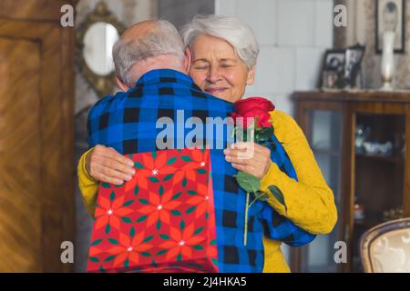 Two people embracing each other. Elderly caucasian couple celebrates birthday, valentines, or anniversary. Friendly grandmother is being hugged by her husband while holding presents. High quality photo Stock Photo