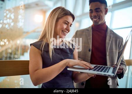 Two young cheerful business people are using a laptop while standing in a pleasant atmosphere in the hallway. Business, people, company Stock Photo