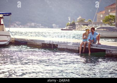 Couple sitting on wooden jetty by sea, having drink and flirting. Boats city and mountains in the background. Love, holiday concept. Stock Photo