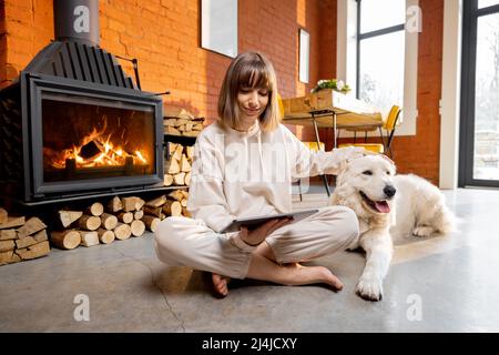 Woman sitting relaxed with her dog on the floor near the fireplace and using digital tablet at home Stock Photo
