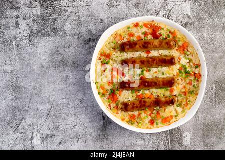 Oven baked rice with sausages and vegetables in a white baking dish on a dark grey background. Top view, flat lay Stock Photo