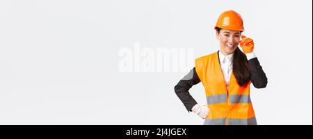 Confident successful asian female chief manager looking proud and smiling at camera, wearing safety helmet, glasses and gloves at construction area or Stock Photo