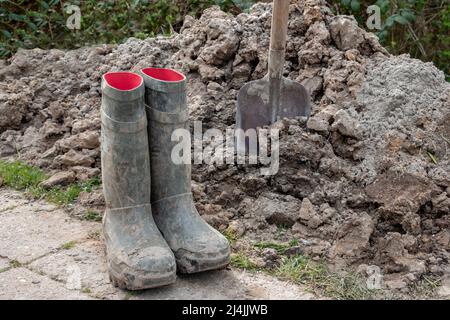 A shovel in a heap of clay soil, after hand-digging a piece of soil and having dirty green boots Stock Photo