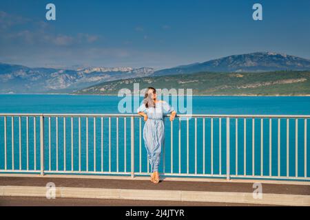The beautiful girl in a blue dress and sunglasses poses on the bridge, the long chestnut hair, happy and smiles, azure water of the lake and slopes of