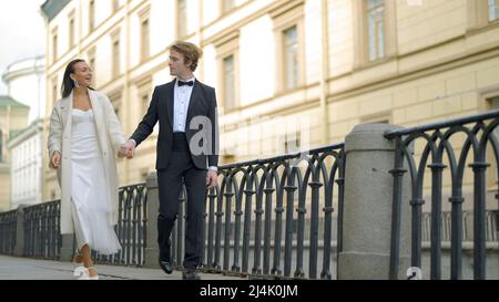 Beautiful newlyweds walking around city. Action. Happy and elegant newlyweds are walking along pavement of city canal. Walking newlyweds in city Stock Photo