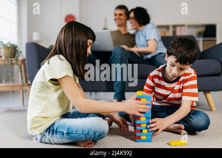 Kids playing on the floor while parents using a laptop in the living room at home. Stock Photo