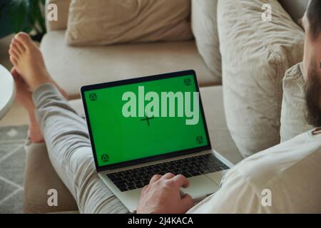 Young man using laptop green screen chroma key, working with touch pad, lying on sofa at home Stock Photo