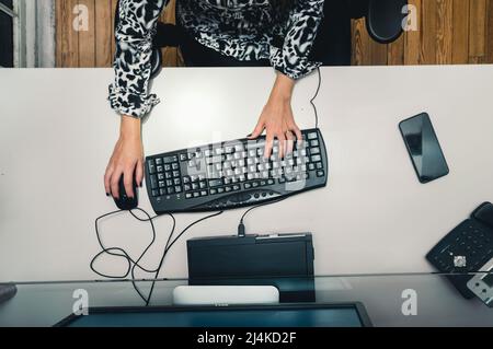 top view unrecognizable young woman sitting at the desk with her hands on the keyboard and mouse, working with the computer in an office programming, Stock Photo