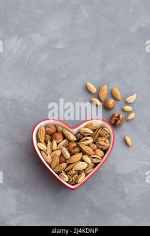 Variety of nuts in red bowl heart shaped on gray background. almonds, walnuts, hazelnuts, pistachios. healthy food for the heart. top view. horizontal Stock Photo