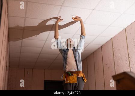 Handsome young builder is twisting the light bulb in. The man is looking up. Stock Photo