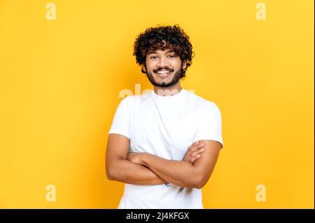Happy positive indian or arabian curly-haired guy in basic white t-shirt, pointing fingers up at empty mockup space for advertising, announcement, standing on isolated orange background, smiling Stock Photo