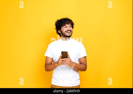 Joyful happy good looking indian or arabian guy holding smartphone in hand, chatting online, browsing internet, looking happily to the side, standing on isolated orange background, smiling Stock Photo