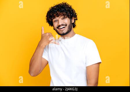 Call me baby. Positive indian or arabian millennial man holding hand near ear, showing telephone sign with fingers, asking for number, standing on isolated orange background, looks at camera, smiling Stock Photo