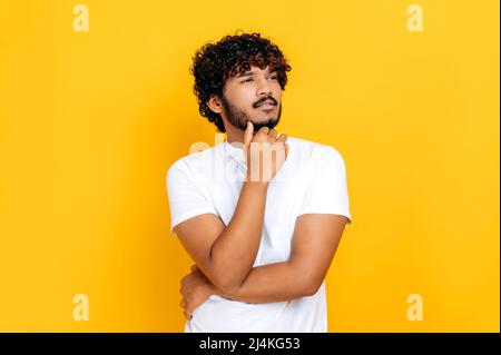 Portrait of a handsome optimistic indian or arabian guy in basic t-shirt, looking pensively to the side at empty space, thinking new ideas, stands on isolated orange background, holds hand near chin Stock Photo