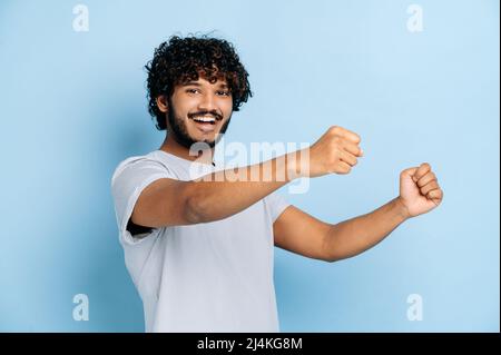Cheerful attractive curly haired indian or arabian guy, wearing t-shirt, holding in hands driving invisible car, imaginary steering wheel, stands on isolated blue background, smiling, looks at camera Stock Photo