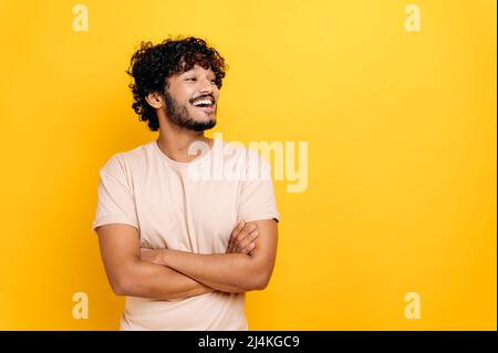 Charismatic attractive arabian or indian curly-haired guy in a t-shirt, freelancer, student, standing over isolated orange background with crossed arms, looking away, smiling happily Stock Photo